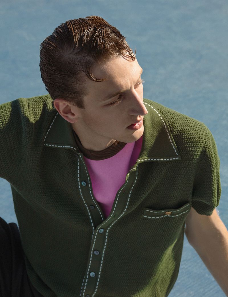 Model Jaume Marti looking away wearing an olive green knit short-sleeve shirt over a pink undershirt against a blue surface.