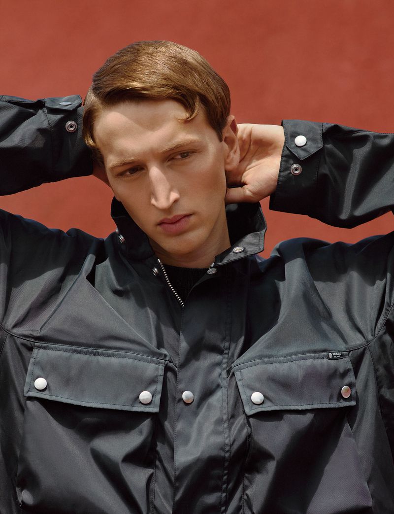 Model Jaume Marti poses with hands behind his head wearing a black utility snap-button jacket against a terracotta background.
