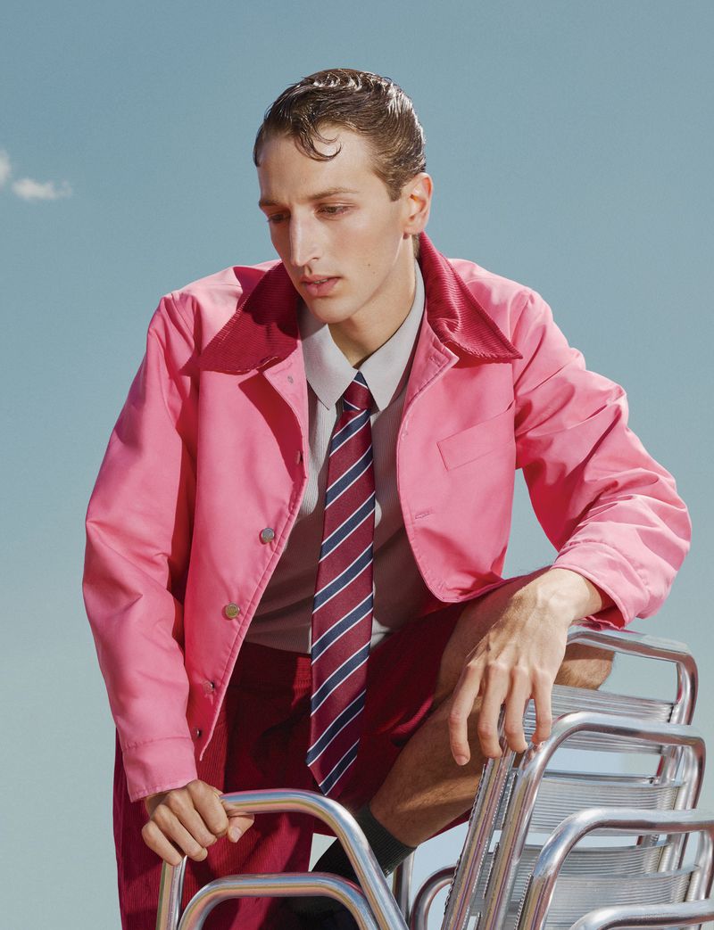 Model Jaume Marti in a bright pink collared jacket and striped tie leaning over stacked metal chairs against a pale blue sky.
