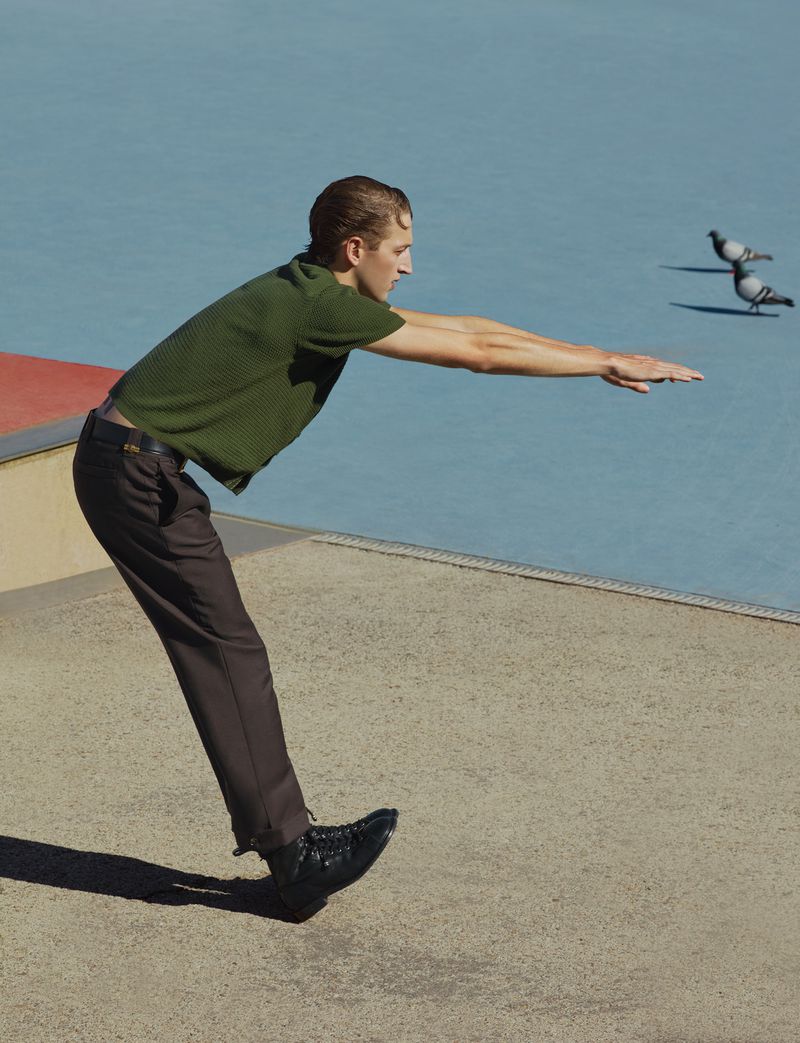 Model Jaume Marti leaning forward with arms extended, wearing an olive green knit shirt and brown trousers on concrete.