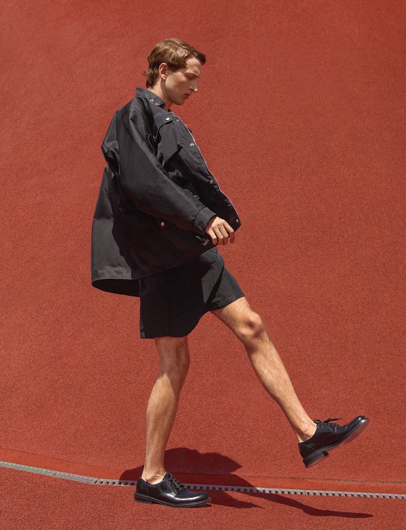 Model Jaume Marti mid-stride in a black jacket, shorts, and leather shoes against a curved terracotta red athletic surface.