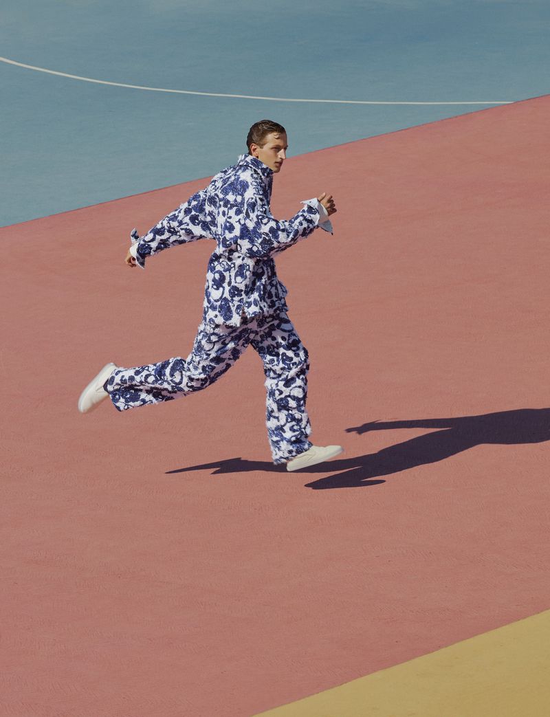 Model Jaume Marti running across a color-blocked outdoor court wearing a blue and white patterned matching suit and white sneakers.