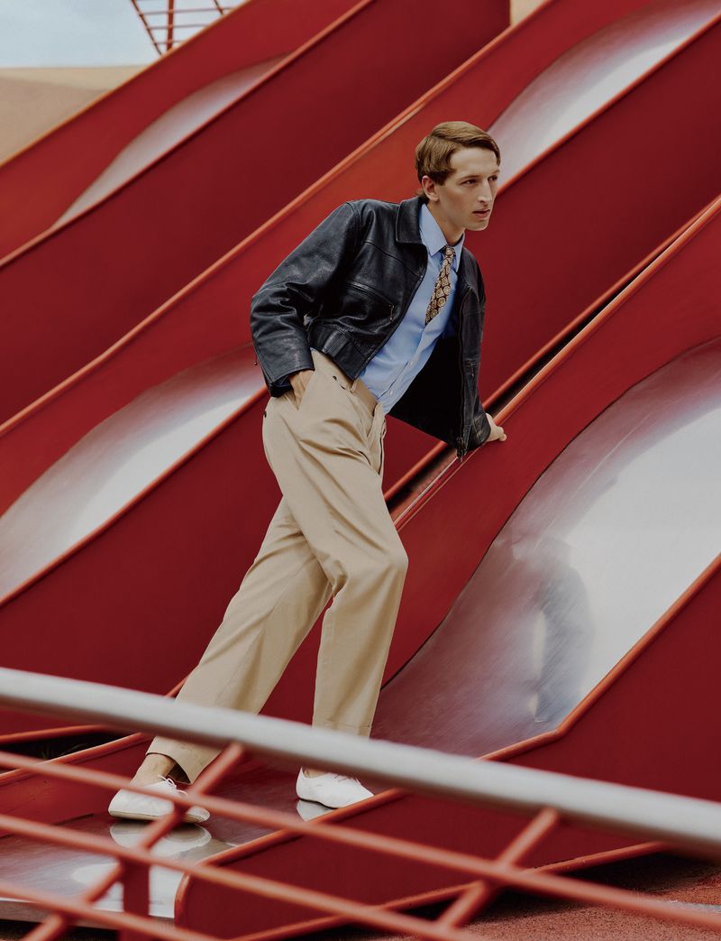 Model Jaume Marti in a black leather jacket and beige trousers walking up a red playground slide structure.