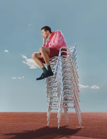 Model Jaume Marti sitting thoughtfully atop a tall stack of silver metal chairs on a red running track under a blue sky.