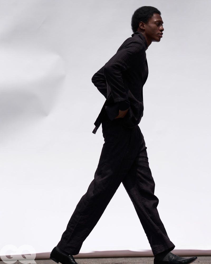 Side profile of a man walking in a black tailored suit and dress shoes across a white seamless studio backdrop.