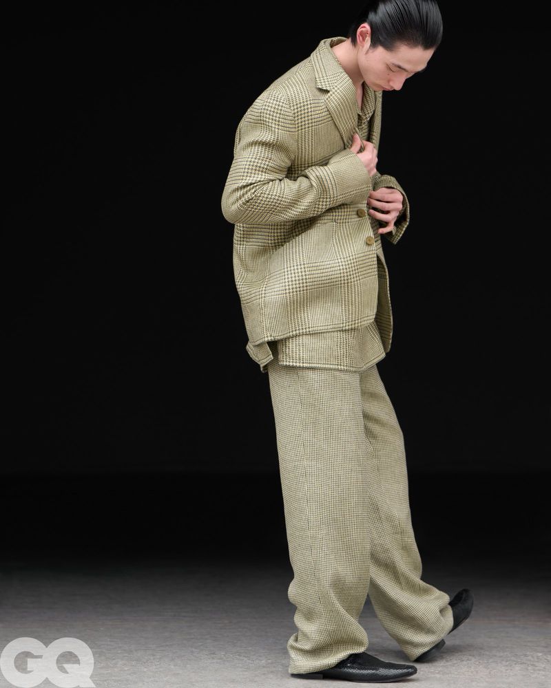 Model Chengwei Dong wearing an oversized olive plaid suit and black dress shoes against a dark backdrop.