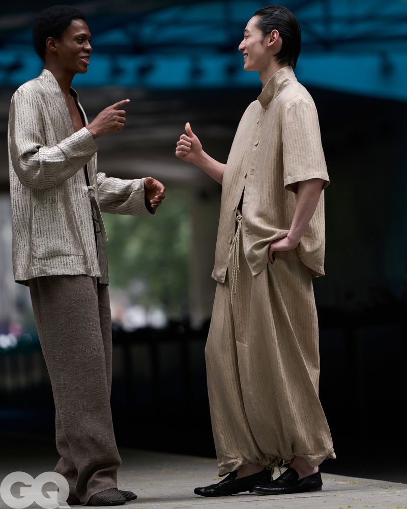 Two male models gesturing and talking in relaxed neutral textured shirts and wide trousers outdoors under an urban structure.