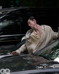 Model Chengwei Dong in a draped beige jacket and striped shirt leaning across dark car hoods for GQ China.