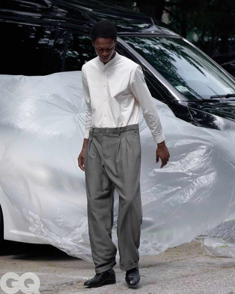 Chengwei Dong walks wearing a white band-collar shirt and grey pleated trousers in front of a car wrapped in plastic sheeting.