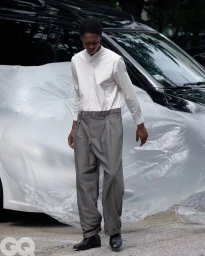Chengwei Dong walks wearing a white band-collar shirt and grey pleated trousers in front of a car wrapped in plastic sheeting.