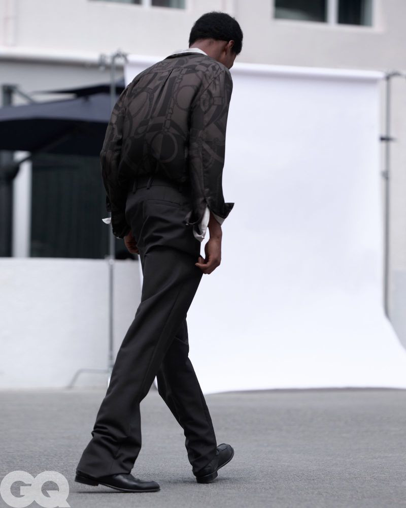 Model Chengwei Dong walking away in a patterned dark jacket and tailored trousers in front of a white backdrop for GQ China.