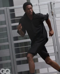 A man running in an all-black collared shirt and matching shorts in front of a light grey slatted metal gate.