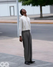 Model wearing a cream band-collar shirt and high-waisted pleated grey trousers standing on a city pavement with head tilted back.