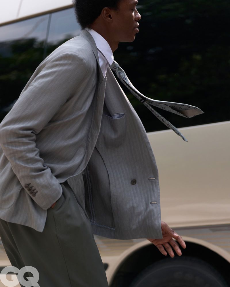 Model Chengwei Dong wearing a grey suit jacket and blowing tie in motion beside a vehicle for GQ China.
