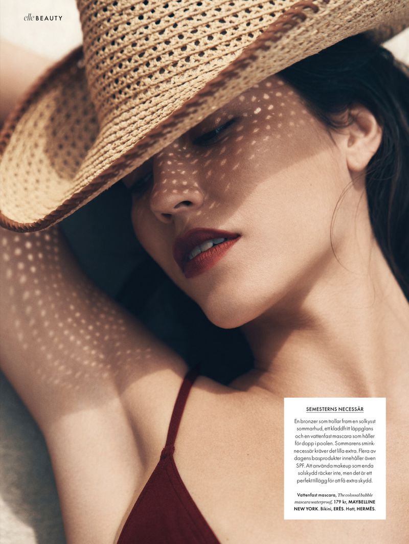 Woman wearing a straw hat in a close-up shot, casting dappled shadows across her face in a beauty advertisement with 'Elle Beauty' logo.