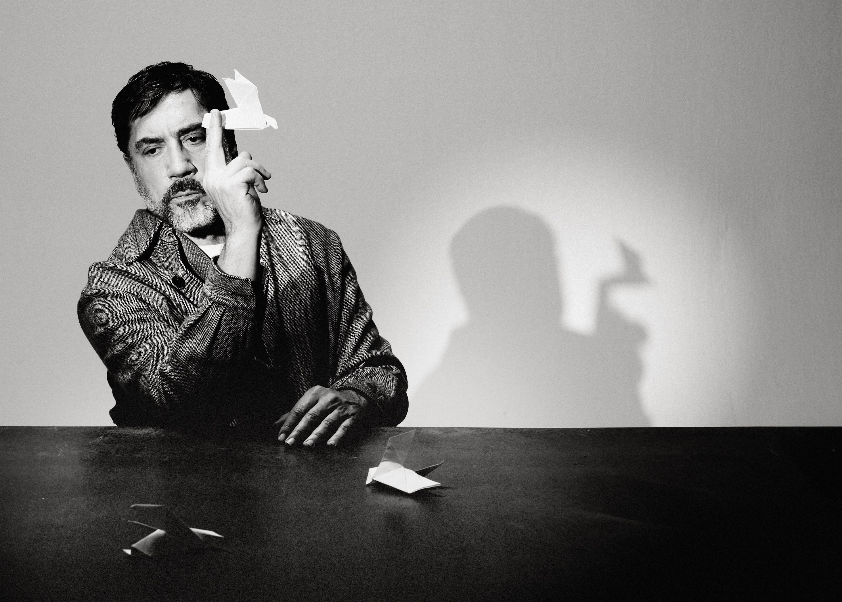 Black and white portrait of Javier Bardem seated at a table balancing a white origami bird on his finger.