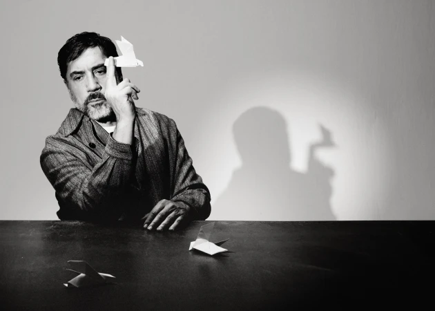Black and white portrait of Javier Bardem seated at a table balancing a white origami bird on his finger.