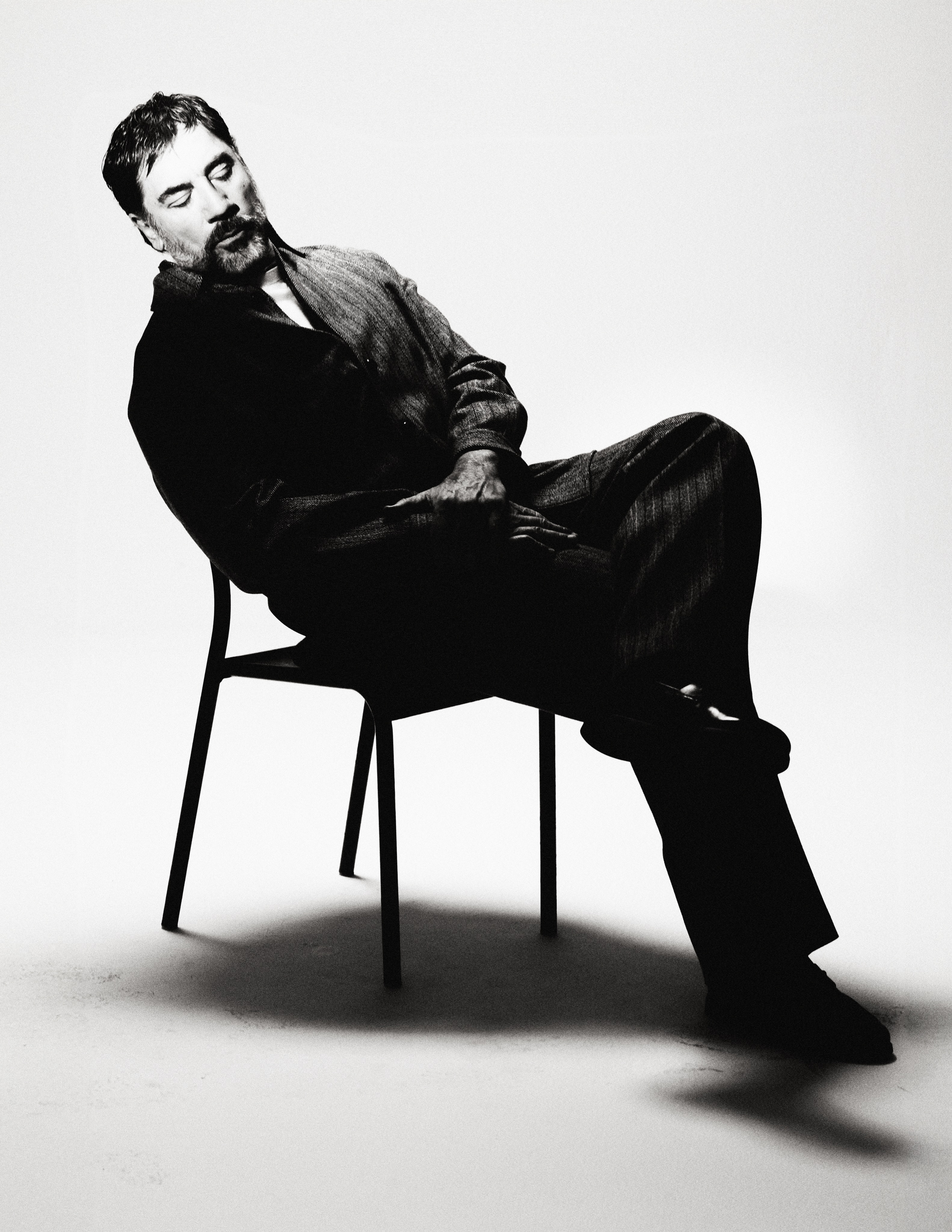 Black and white portrait of actor Javier Bardem seated back on a chair with eyes closed against a stark white studio backdrop.