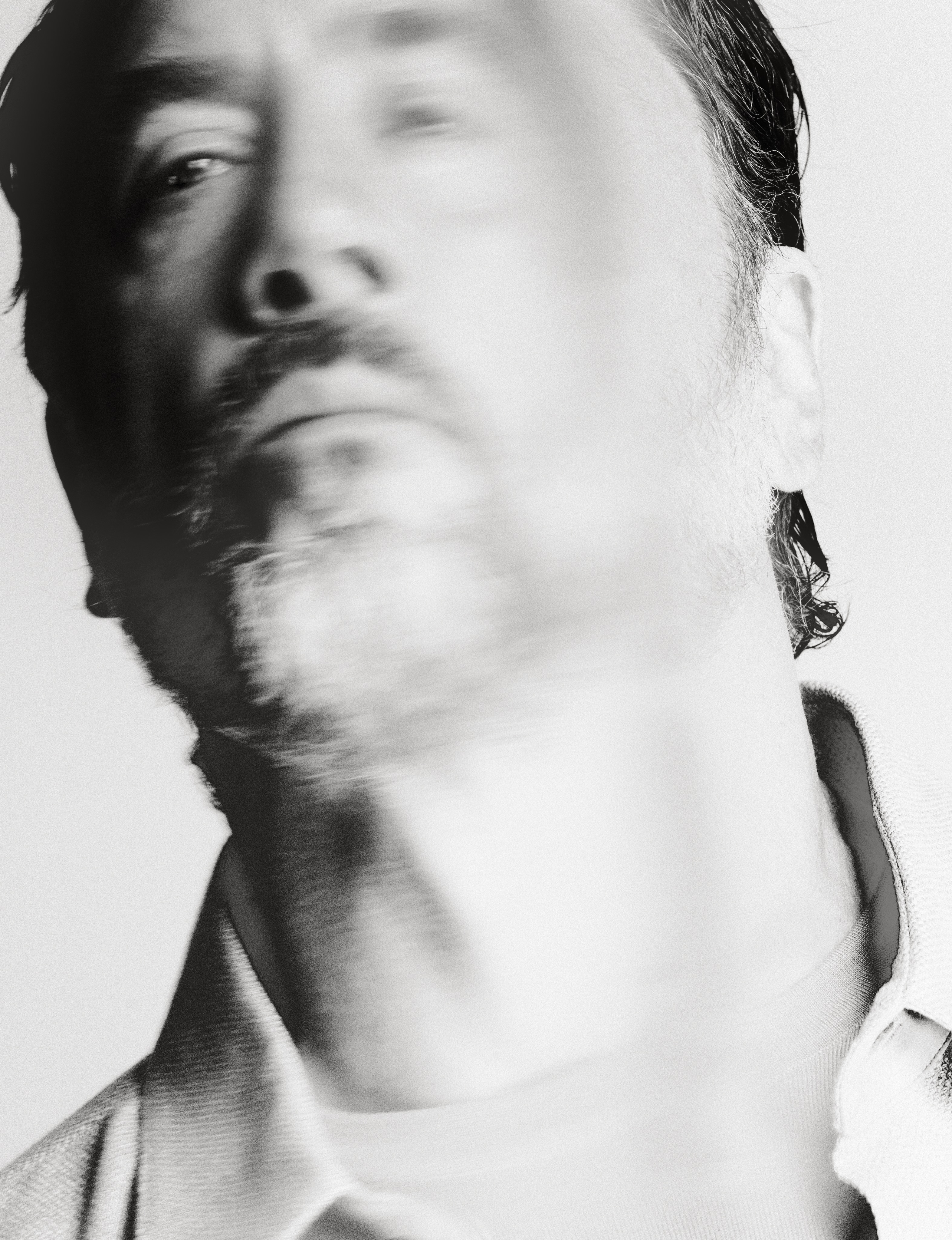 Black and white close-up portrait of actor Javier Bardem with motion blur looking upward against a pale background.