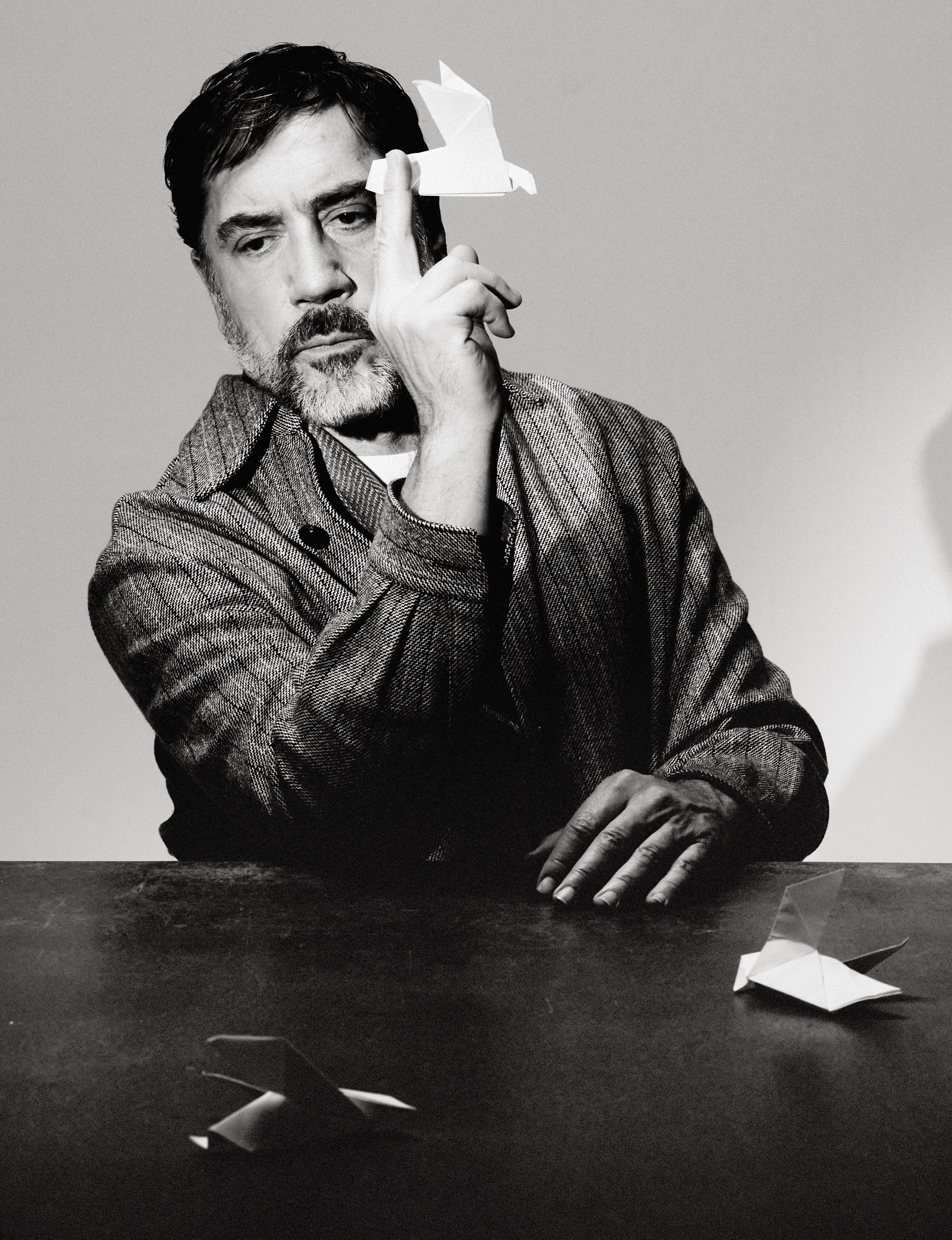 Javier Bardem balances a white paper origami bird on his index finger while seated at a dark table wearing a textured jacket.