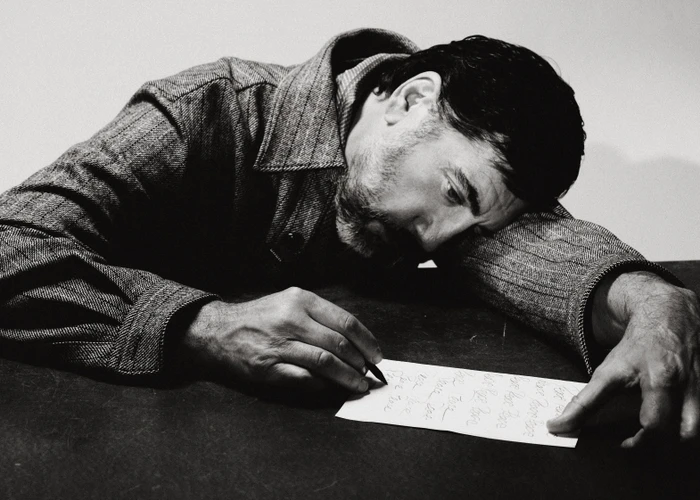 Javier Bardem leaning over a table in a textured coat, writing on a piece of paper in a black and white editorial shoot.