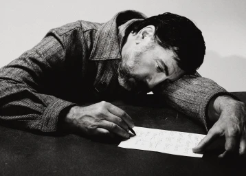 Javier Bardem leaning over a table in a textured coat, writing on a piece of paper in a black and white editorial shoot.