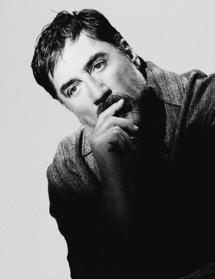 Black and white portrait of actor Javier Bardem resting his hand on his chin against a plain light studio backdrop.
