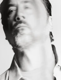 Monochrome portrait of actor Javier Bardem with intentional motion blur, wearing a light collared shirt against a white backdrop.