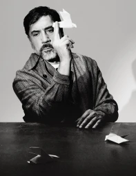 Javier Bardem balances a white paper origami bird on his index finger while seated at a dark table wearing a textured jacket.