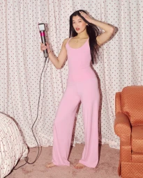 Woman in a pink tank top and matching wide-leg pants holding a hair styler before patterned curtains and an armchair.
