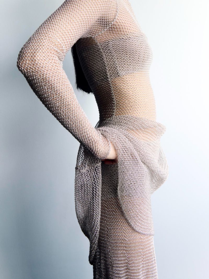 Side view of an adult female wearing a sheer, patterned dress with long sleeves against a light background in a studio setting.