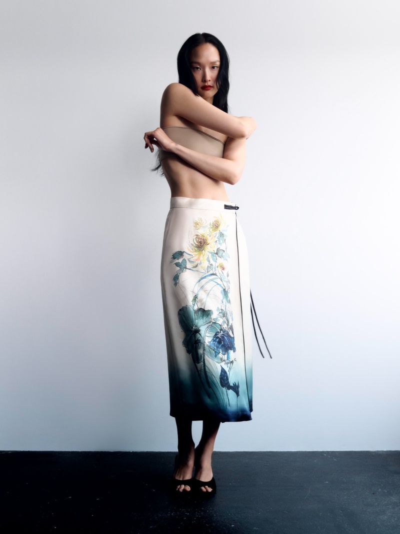 Fashion photo of a woman wearing a beige tube top and a long skirt with floral designs, posing in front of a plain white wall.