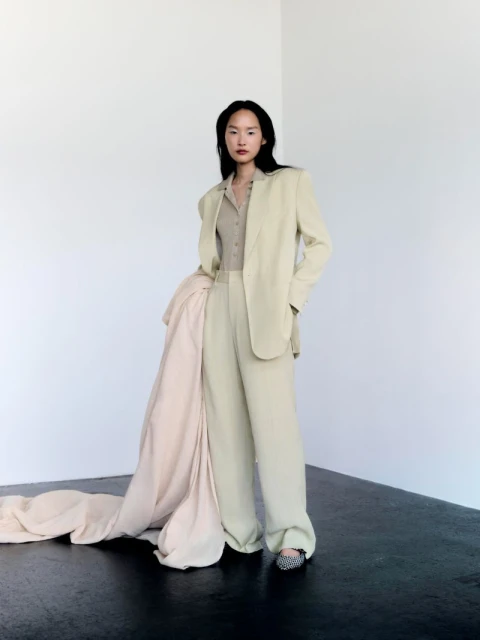 Elegant woman in a light-beige suit with wide-leg pants and flowing jacket in a minimalist studio setting, showcasing modern fashion.