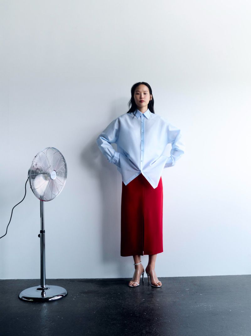 An Asian woman in a blue button-down shirt and red midi skirt stands in a photo studio next to a silver standing fan against a white wall.