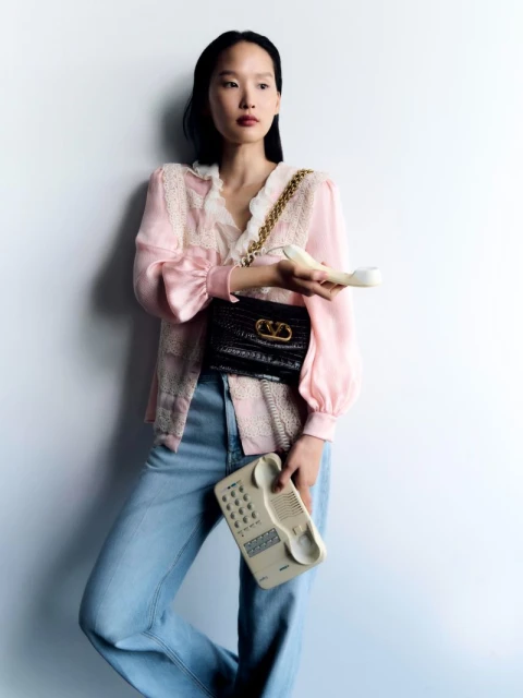 Elegant Asian woman in pink blouse and jeans holds a vintage phone and a black Valentino purse against a plain white backdrop.