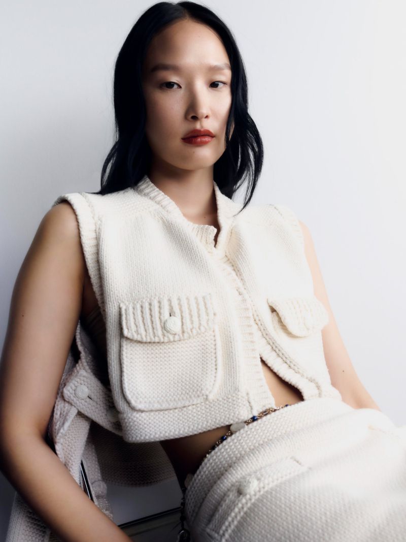 Woman in an ivory knit vest and skirt posing against a white wall. The fashion shot has diffused lighting and neutral colors.