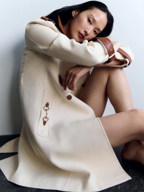 An Asian woman in a light beige trench coat poses indoors, seated on the floor, with a bright, neutral backdrop in a fashion setting.