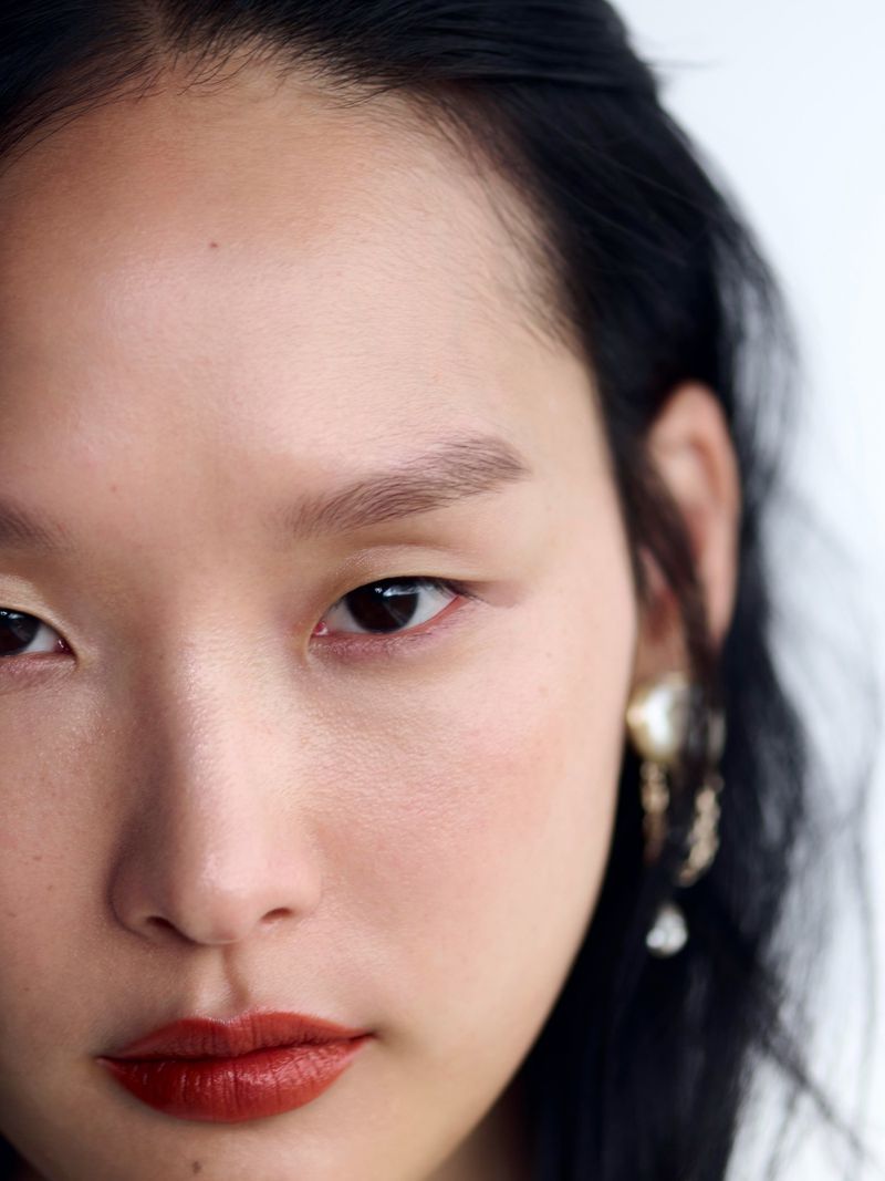 Close-up portrait of a young Asian woman with dark hair and red lipstick, adorned with ornate gold earrings, against a bright white background.