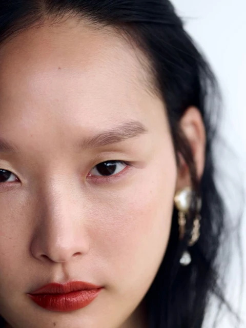 Close-up portrait of a young Asian woman with dark hair and red lipstick, adorned with ornate gold earrings, against a bright white background.