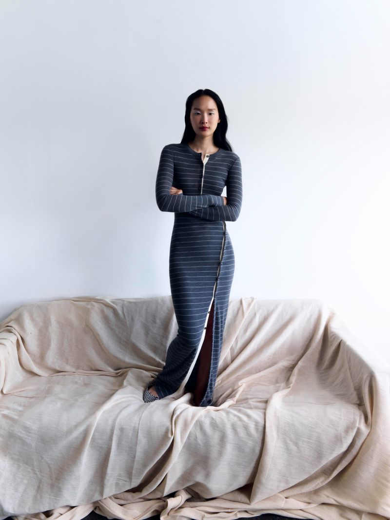 Elegant woman poses in a gray striped maxi dress with a front zipper, standing on a draped couch against a plain white wall.