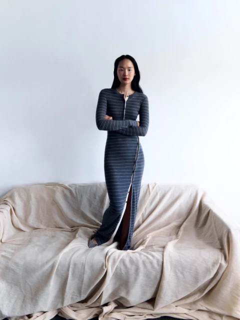 Elegant woman poses in a gray striped maxi dress with a front zipper, standing on a draped couch against a plain white wall.