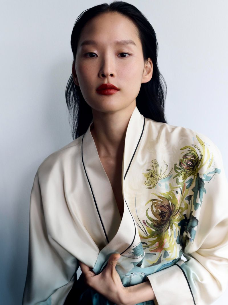 Adult woman with black hair, wearing a floral patterned kimono and red lipstick, poses in front of a white background in a portrait shot.