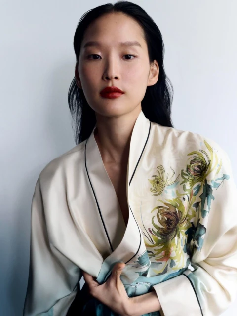 Adult woman with black hair, wearing a floral patterned kimono and red lipstick, poses in front of a white background in a portrait shot.
