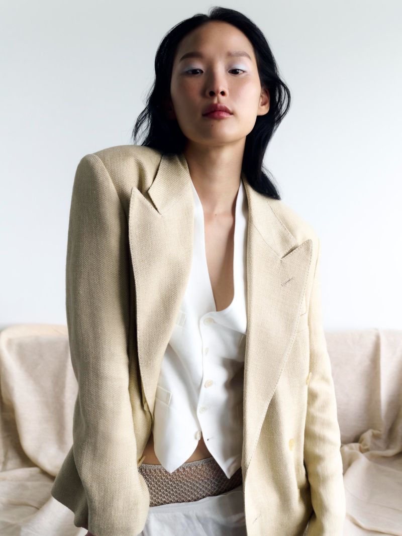 Asian woman in a beige blazer and white vest poses against a draped fabric backdrop with bright eyes and dark hair in a fashion portrait.