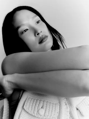 Close-up monochrome portrait of an Asian model with dark hair and folded arms wearing a textured knit top.