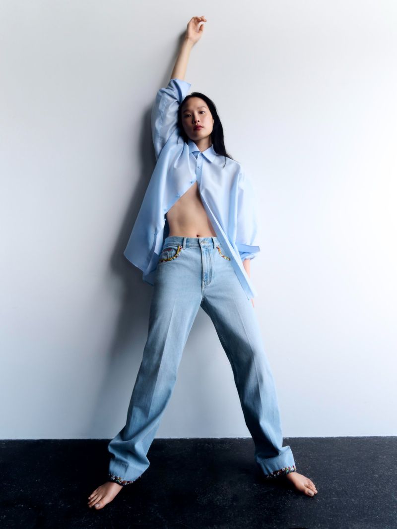 Model in open blue shirt and light blue jeans posing against a white studio wall with her arm raised