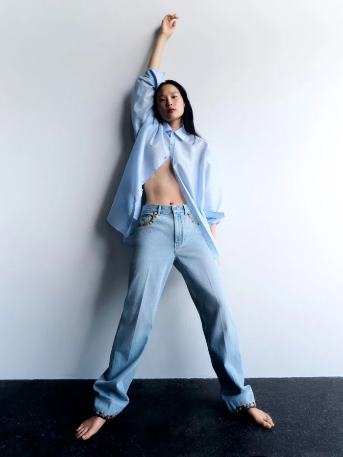 Model in open blue shirt and light blue jeans posing against a white studio wall with her arm raised