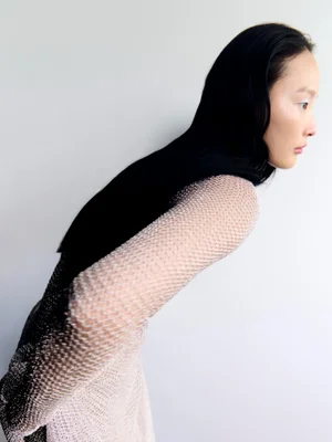 Model in mesh long sleeve top leaning forward against a plain white background