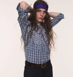 Person with long wavy hair wearing a blue checked button-up shirt, dark trousers, and a purple paper crown against a white backdrop.