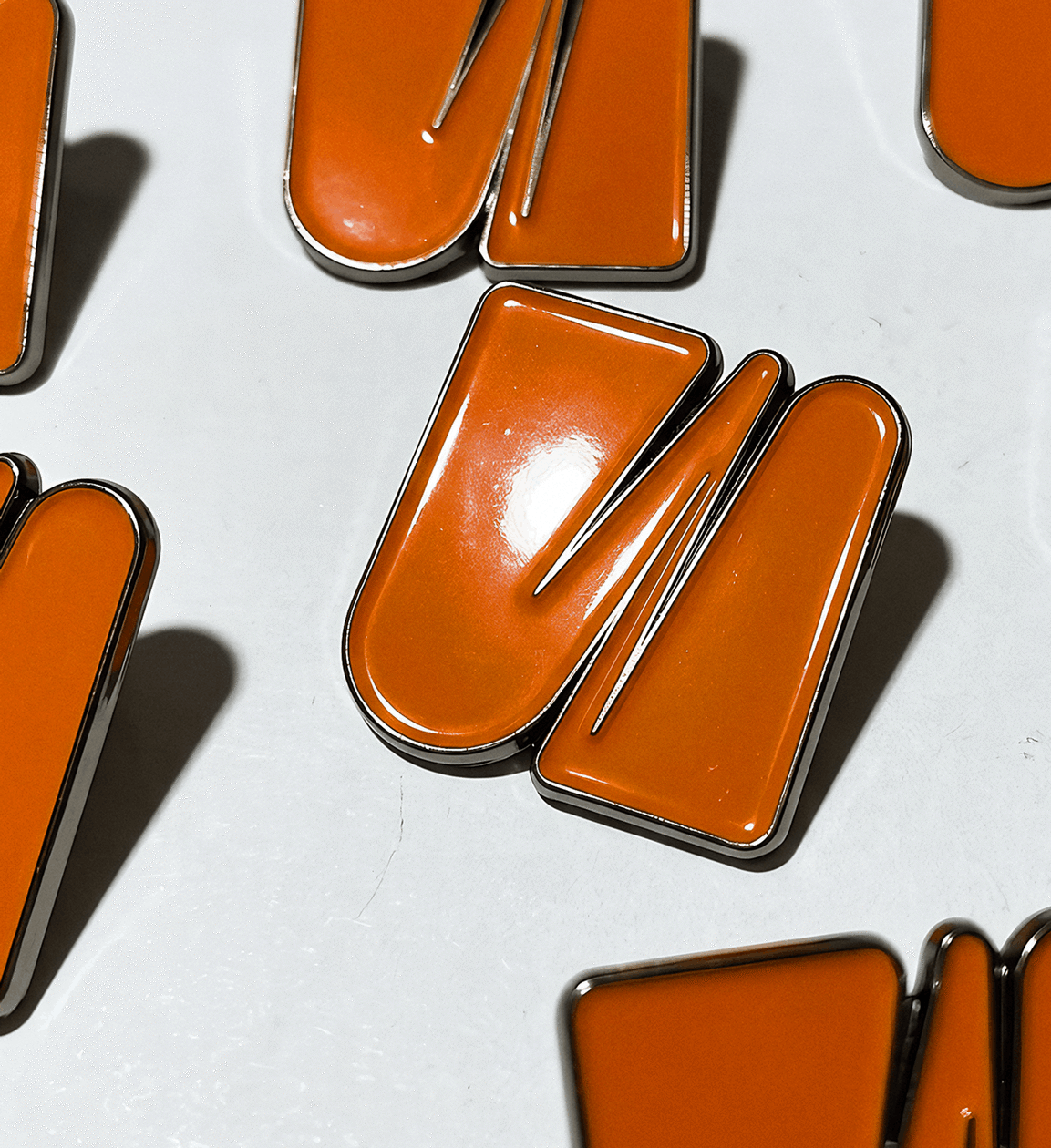 Glossy orange enamel pins with silver metal edges arranged across a plain white surface casting distinct shadows.